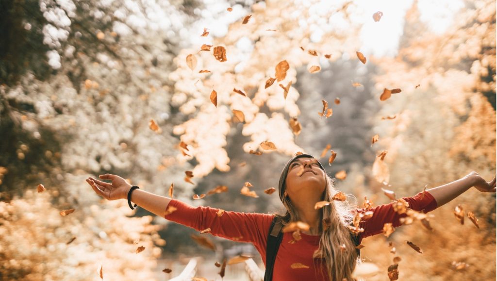 On a sunny day outside, a woman looks up with outstretched arms as autumn leaves fall around her.