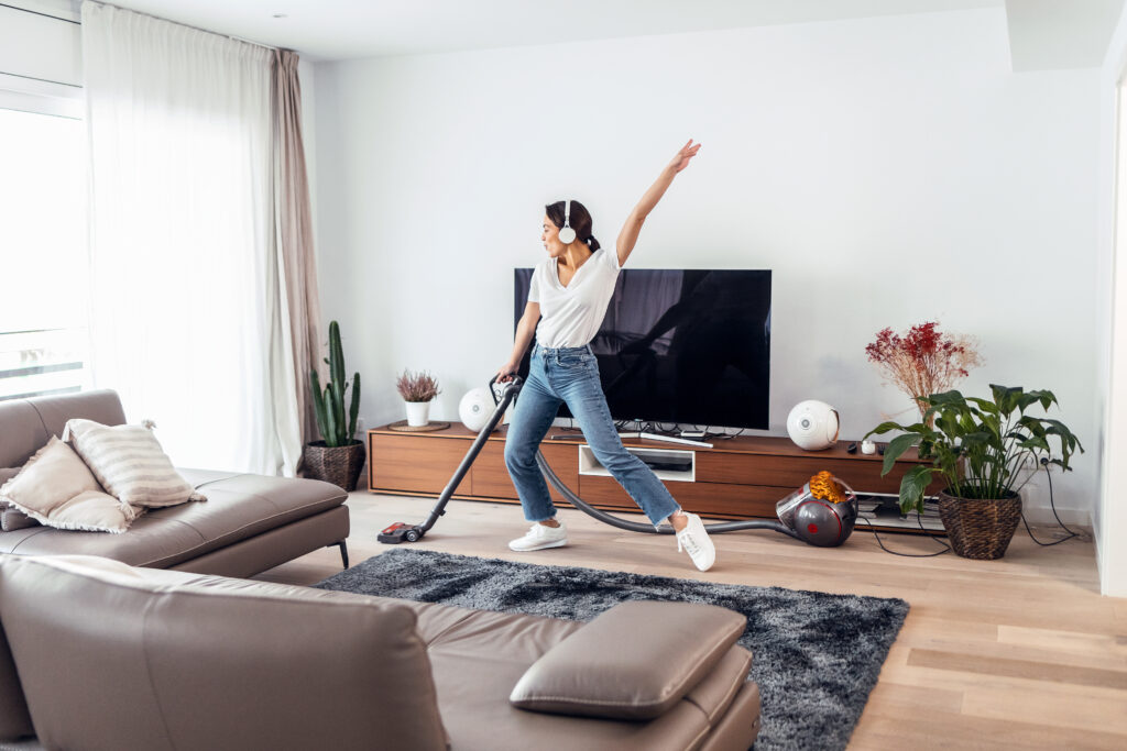 A woman dances to music on her headphones as she vacuums the living room.