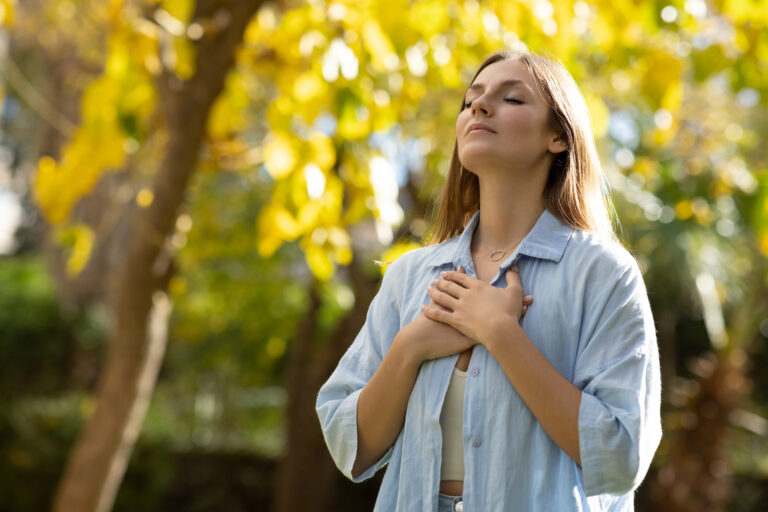 Standing among trees, a woman stands with a calm expression, her eyes closed and her hands on her heart.