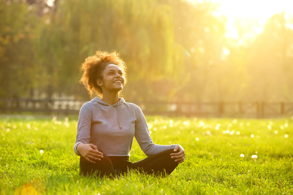 A smiling woman sits in a relaxed yoga pose on the grass on a sunny day.
