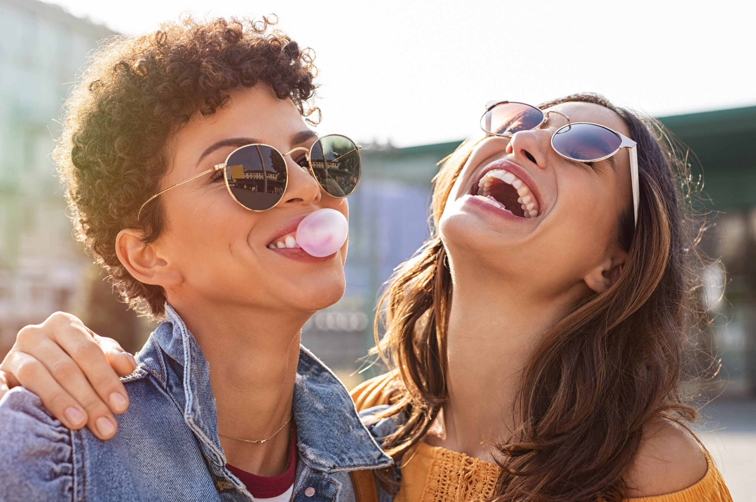 Outdoors in the sun, two women in jean jackets and sunglasses are laughing together while one blows a bubble with bubblegum.