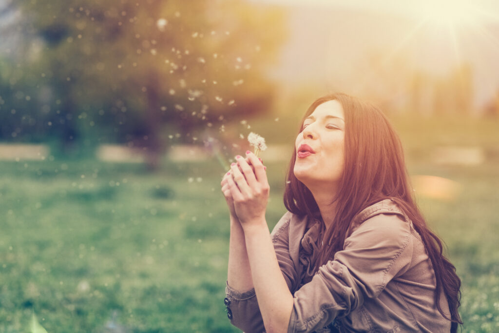 Outside on a lawn, a woman sits on the grass and blows a dandelion puff, sending the fluffy seeds flying away.