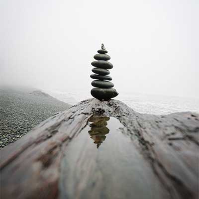 Against a misty horizon on a black sand beach, a stack of smooth stones sits at the far end of a tidal pool and is reflected in the water.