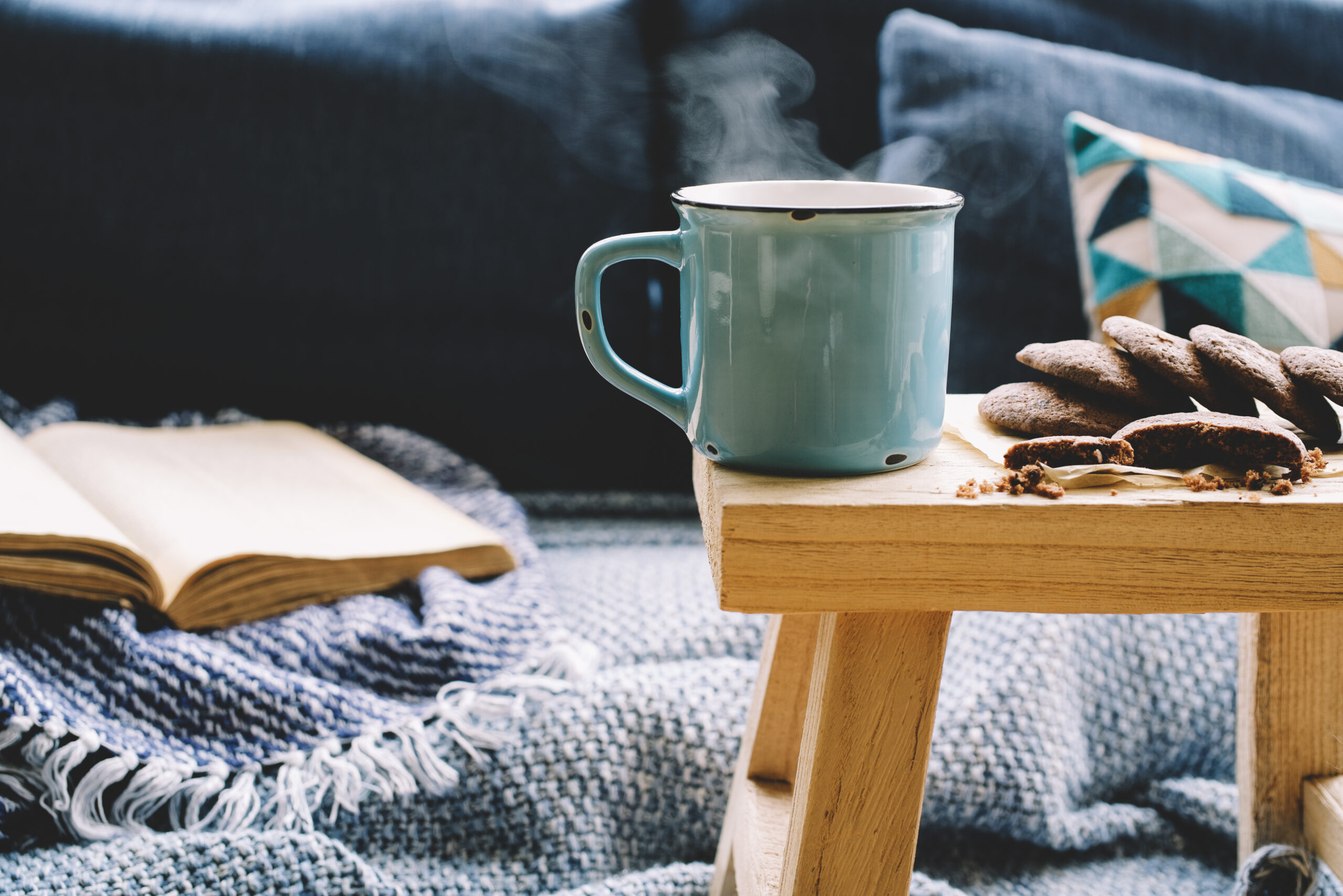 A cup of coffee sits on the edge of a wooden bed tray. On the bed is a cozy blanket and a book.