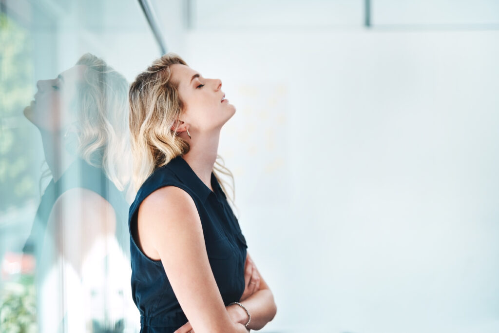 In an office, a woman leans back against the glass wall, her arms folded and her eyes closed, and a stressed expression on her face.