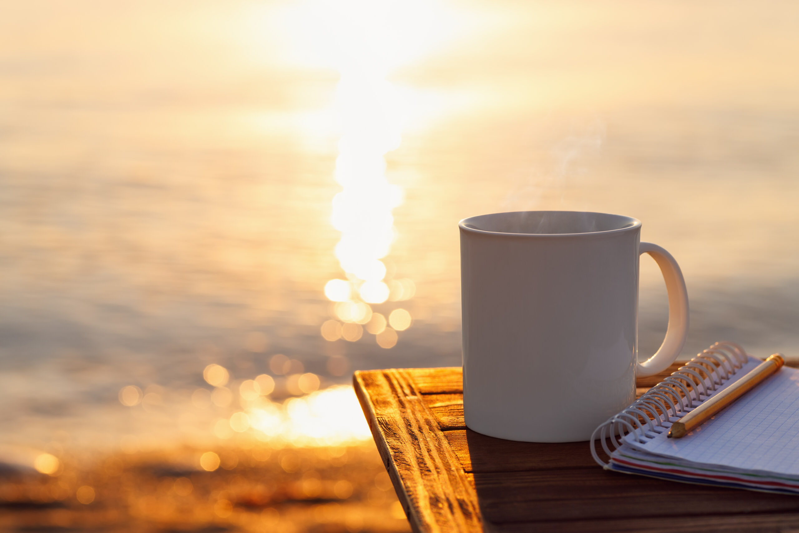 Against a calm ocean sunrise in the background, a white coffee cup sits on a wood table.
