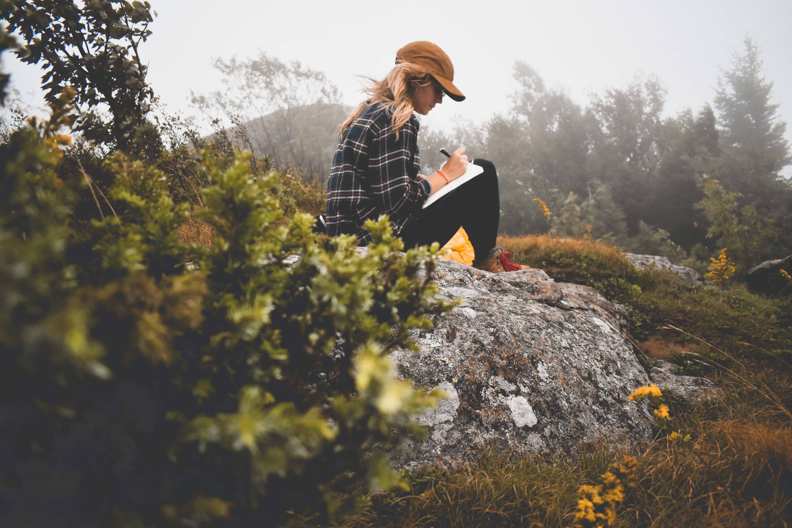 A woman out in nature sits on top of a rock journaling.