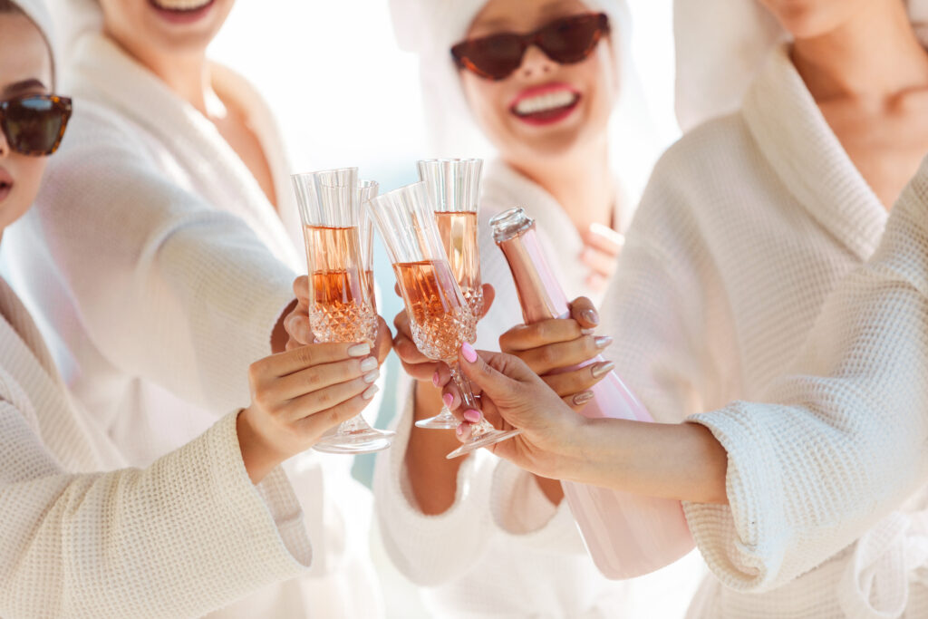 A group of women in sunglasses, spa robes, and hair towels clink champagne glasses together in a toast.