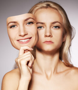 A woman faces the camera with a somber expression while holding a mask of her own smiling face which she has just moved to the side to show her real expression underneath.