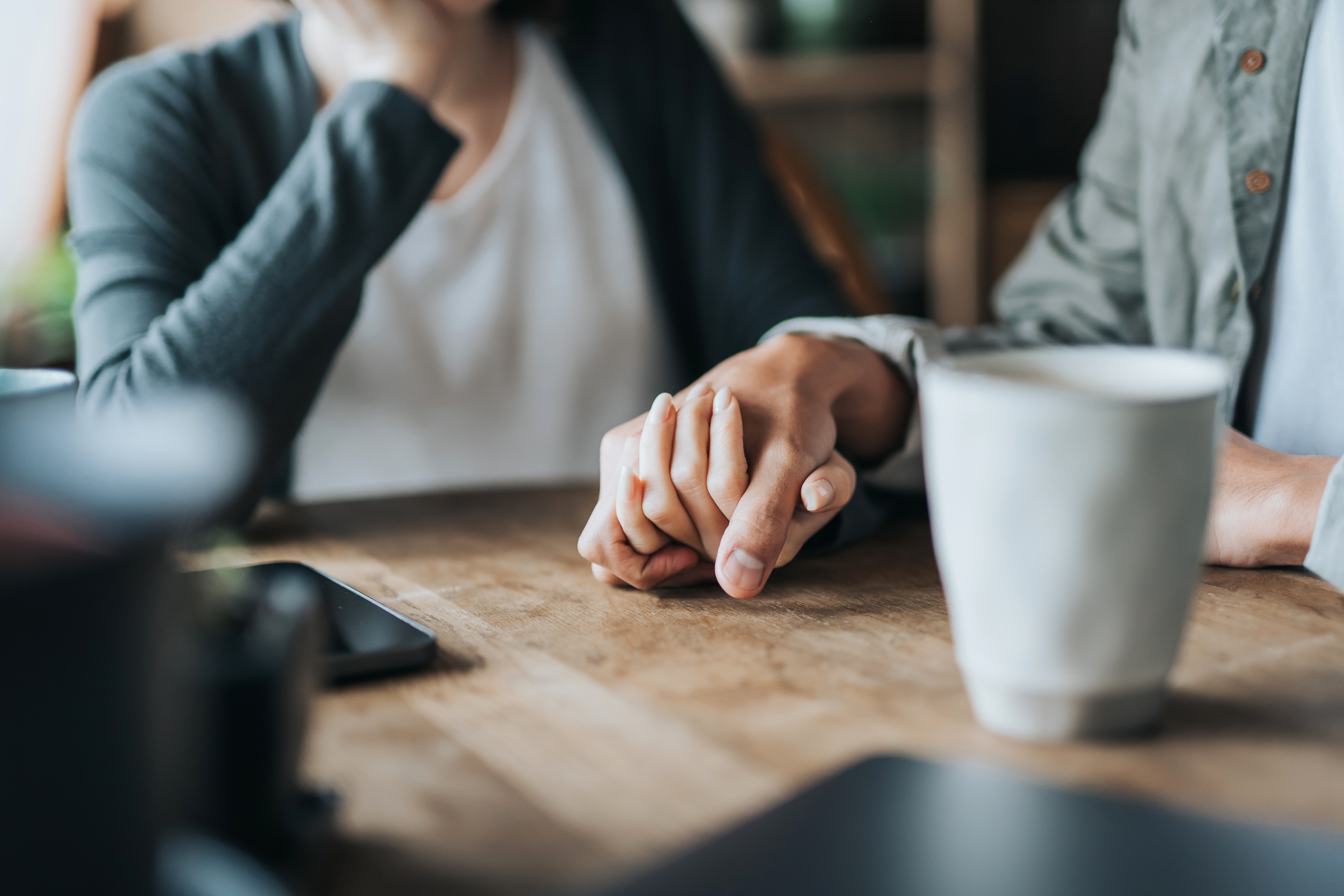 On a wooden table with some white coffee cups, the view is of two people holding hands to support each other while they're worried.