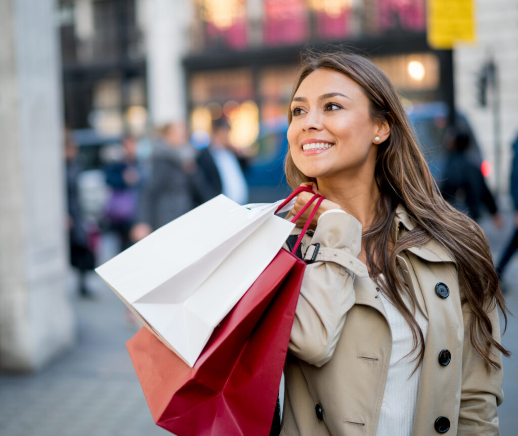 A woman looks around happily as she walks downtown with bags from various places she's been shopping.