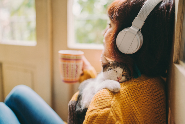 A woman listens to music on her headphones and enjoys a cup of tea as her small cat sleeps on her shoulder.