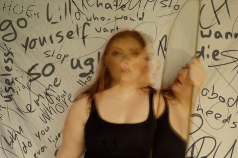 A double exposure of a woman looking toward the camera overlaid with a side profile of her looking into a mirror. Behind her is a background with handwritten cruel self talk.
