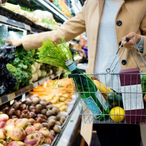 A woman does grocery shopping - she carries a metal shopping basket through the produce aisle and has added some vegetables.